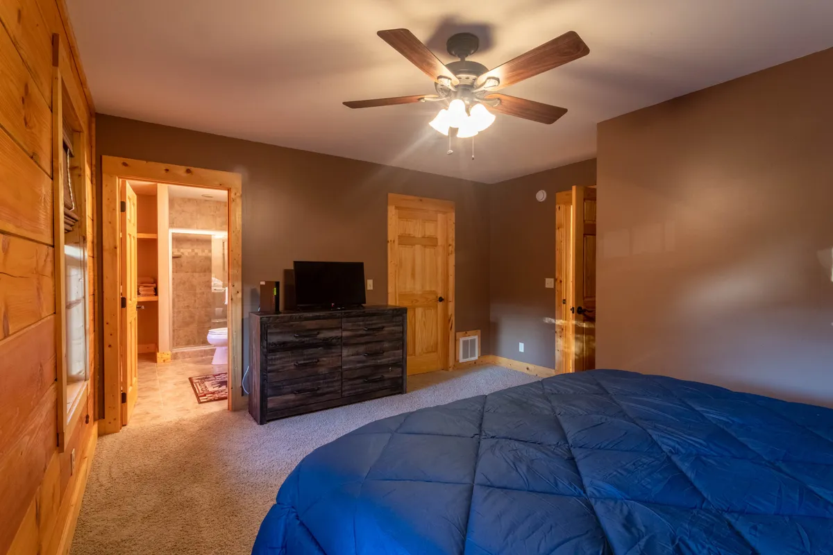 A serene bedroom with a bed featuring blue bedding, wooden furniture, a ceiling fan, and a TV positioned for comfortable viewing from bed. The atmosphere is homey and restful, suggesting both relaxation and the availability of modern comforts like streaming TV.