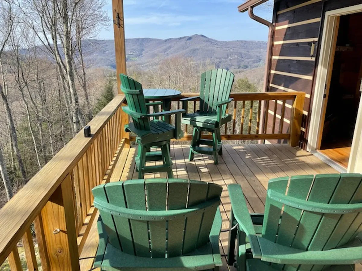 This exterior image features green Adirondack chairs arranged around a small round table on a deck, with a beautiful mountain view in the background. The scene is tranquil, inviting guests to relax and take in the natural beauty of the surrounding Blue Ridge mountains.