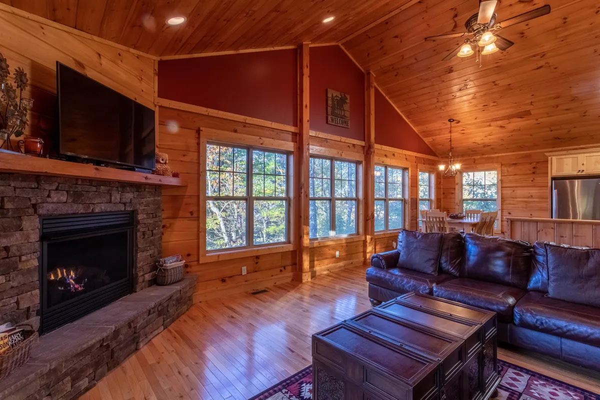 An expansive open-plan living and dining area with a prominent stone fireplace, comfortable sofa, wooden ceiling, and a large window that frames the mountain scenery. The visible dining area invites gatherings for meals or games, while the overall setting is perfect for sharing stories or relaxing nights in.