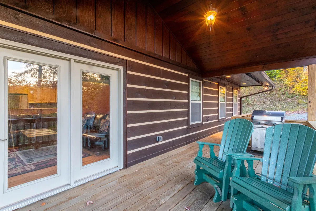 This image showcases the deck area with green Adirondack chairs, a wooden deck, and a visible outdoor grill positioned beside sliding glass doors. The setup suggests a casual, social atmosphere perfect for barbecues, morning coffee, or unwinding in the fresh mountain air.