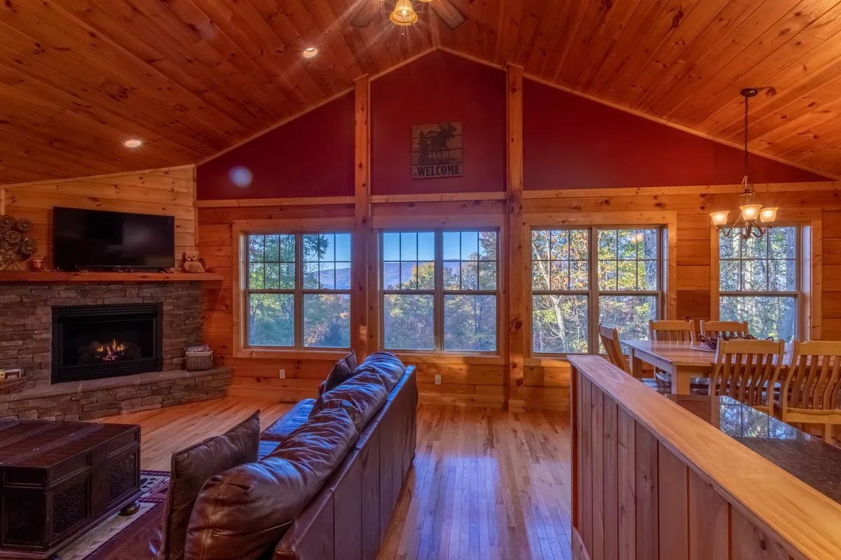 A cozy living room with large windows facing outdoors, a rustic stone fireplace, a wooden dining table, and a plush leather sofa. The room feels warm and welcoming, perfect for coming together after a day outside.