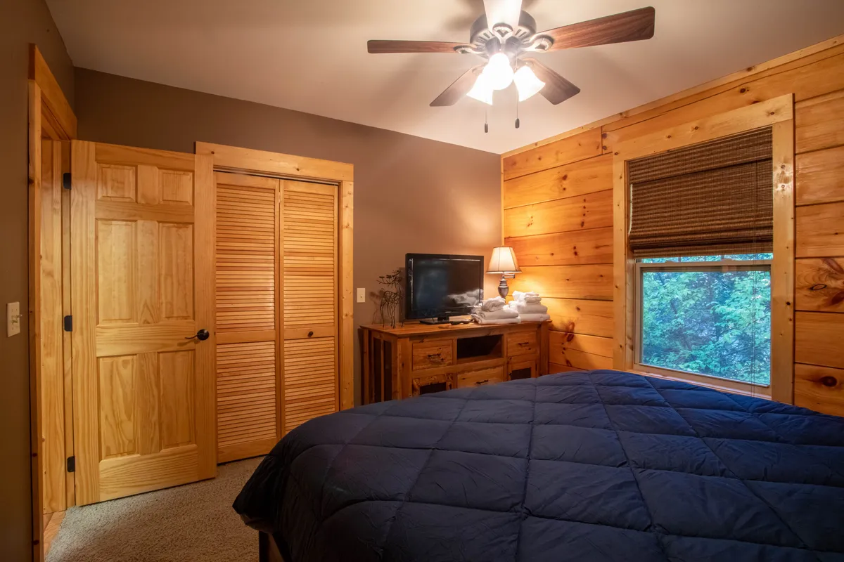 A warm and inviting bedroom featuring a comfortable bed, wooden dresser, and a window that allows natural sunlight to fill the room. The window frames a scenic outdoor view, hinting at the cabin’s connection to its mountain surroundings.