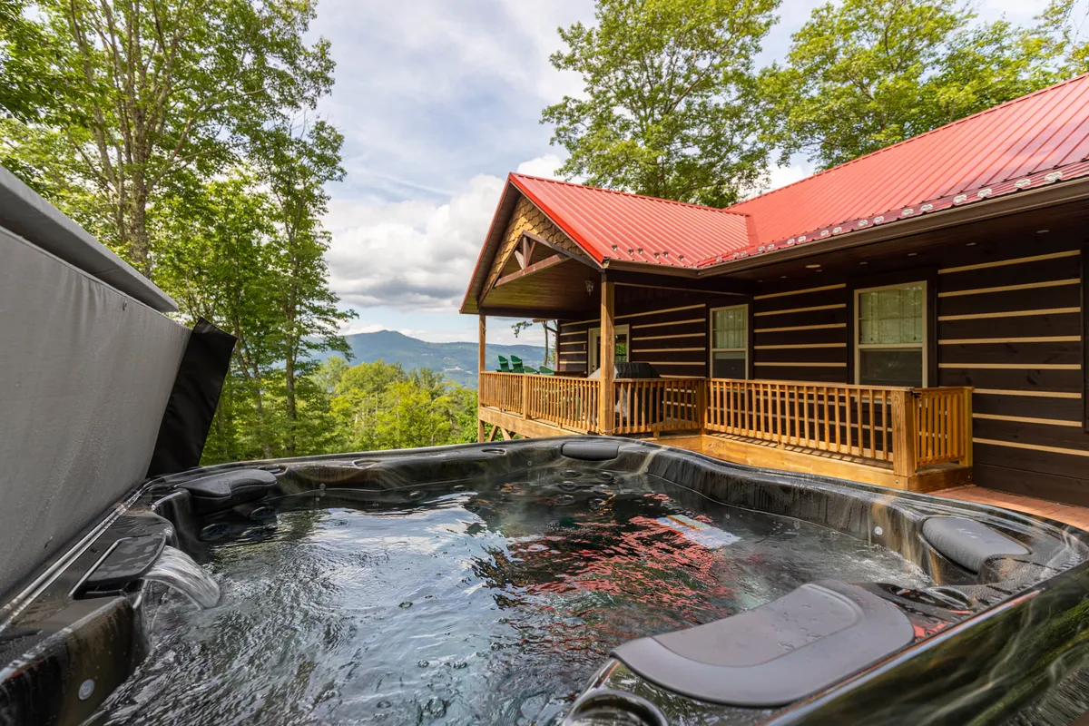 The image shows a charming log cabin nestled in the mountains of Banner Elk, North Carolina. A steaming hot tub sits on the deck, perfectly positioned to take in panoramic mountain views. The setting is serene, with soft evening light suggesting the perfect spot for sunset soaking, and a rustic porch invites guests to relax and unwind.