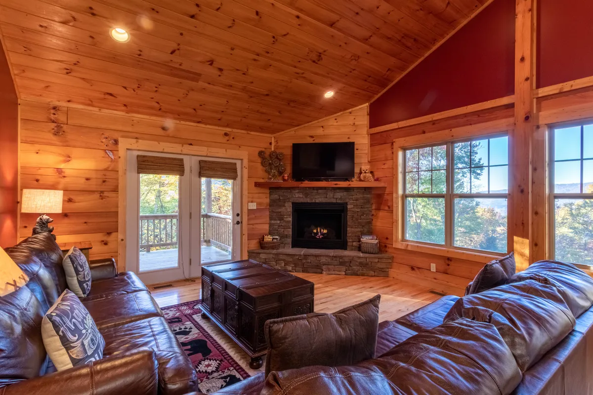 A cozy living room featuring a stone fireplace as the centerpiece, flanked by large windows offering views of the outdoors. The space is anchored by a comfortable sofa and a decorative table, all inviting guests to relax by the fire, gather with family, or enjoy entertainment on the TV, with natural light streaming in to enhance the warm, welcoming ambiance.