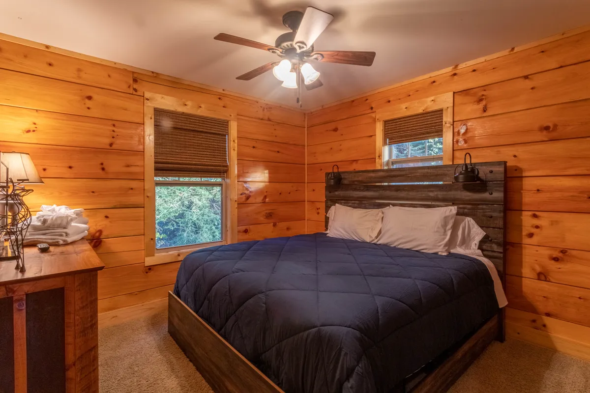 This image features a cozy bedroom with wood-paneled walls, a comfortable bed adorned with a navy comforter, and a nightstand with a lamp. The rustic touches and soft lighting create a welcoming space ideal for relaxation and quiet moments.