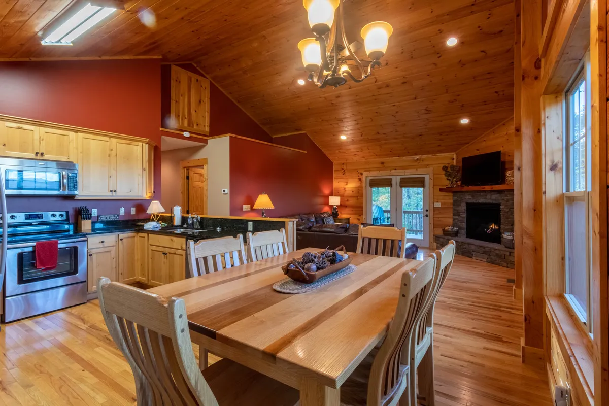 A wide-angle view of the open-concept living space, showing the dining table, kitchen area, fireplace, and living area with large windows letting in natural light. The room features warm wooden finishes, rustic beams, and comfortable furnishings arranged for gatherings. The fireplace anchors the space, and the layout emphasizes flow between kitchen, dining, and living zones.