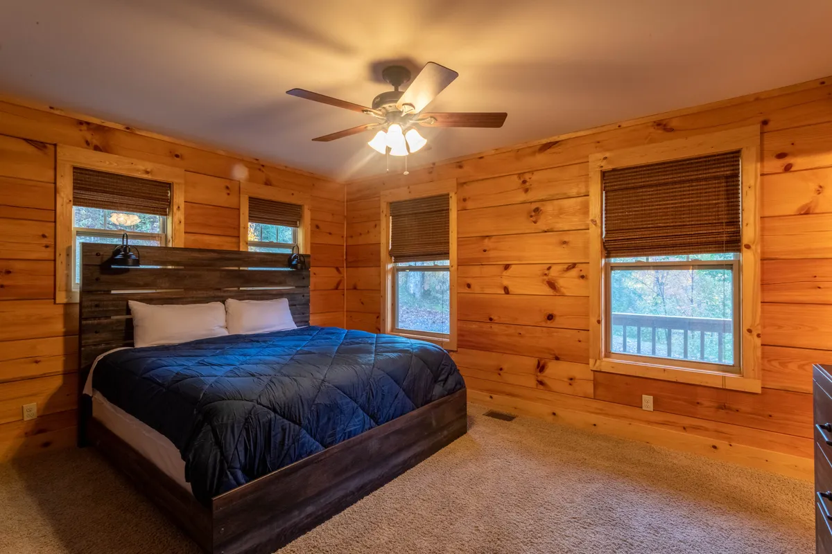 The image depicts a warmly inviting bedroom with wooden walls, a comfortable bed, and large windows offering tranquil nature views. Soft natural light fills the space, creating an atmosphere where guests can relax, read, or simply enjoy the peaceful surroundings.